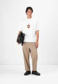 Man met een wit graphic t-shirt, beige broek, zwarte schoenen, die een zwart leren tas vasthoudt tegen een effen witte achtergrond.