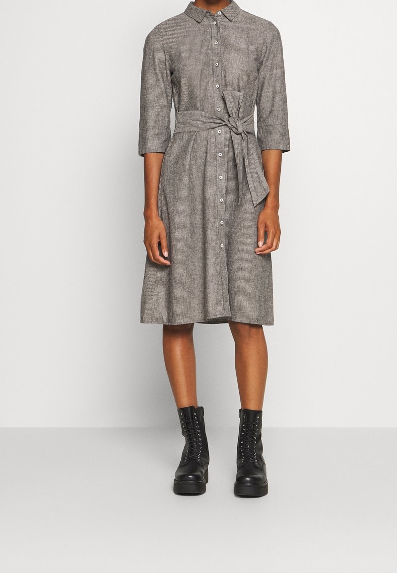 Femme portant une robe grise boutonnée mi-genoux avec une ceinture nouée à la taille et des bottines noires à lacets, debout devant un fond blanc uni.
