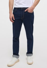Blugi din denim de culoare albastră închisă, cu croială dreaptă, cu cusături portocalii, un nasture frontal și manșete răsucite deasupra unor tenisi albi cu glezna înaltă.
