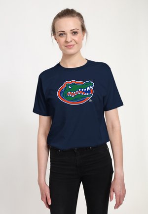 Femme à la peau claire et aux cheveux blonds portant un t-shirt bleu marine avec un logo alligator coloré et un pantalon noir sur un fond uni.