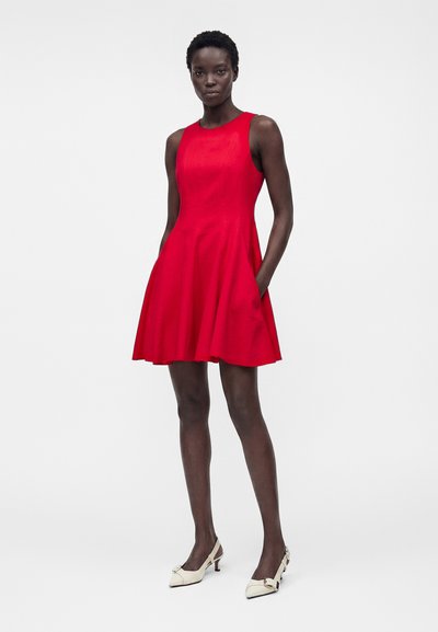 Γυναίκα που στέκεται, φοράει κόκκινο αμάνικο φόρεμα μέχρι το γόνατο με φουσκωτή φούστα και λευκά μυτερά παπούτσια με χαμηλό τακούνι σε απλό φόντο.