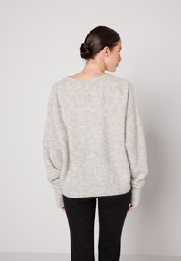 Ženska s temnimi lasmi v nizkem figu, oblečena v svetlo sivi puhasti pulover in črne hlače, obrnjena stran od gledalca na enobarvnem svetlem ozadju.