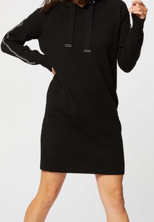 Femme portant une robe à capuche en maille noire à manches longues avec des poches latérales et des détails à rayures argentées sur les manches.