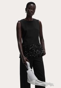 Top negro sin mangas con un dobladillo decorado con lentejuelas, combinado con pantalones negros. La modelo sostiene un bolso ligero gris texturizado con flecos.