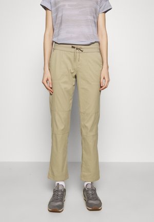 Person trägt ein helllila kurzärmliges Oberteil, beige Hose mit Kordelzug, weiße Socken und graue Turnschuhe und steht vor einem einfarbigen Hintergrund.