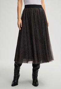 Mulher a usar uma saia midi de malha preta com padrão e cintura elástica, combinada com botas de couro pretas até ao joelho, de pé contra um fundo simples.