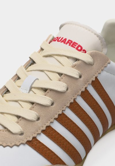 Κοντινό πλάνο άσπρου αθλητικού παπουτσιού με μπεζ σουέτ περιθώριο, εκρού κορδόνια και πέντε καφέ δερμάτινες ρίγες κατά μήκος της πλευράς· μερικά τμήματα κόκκινου κειμένου ορατά στη γλώσσα.