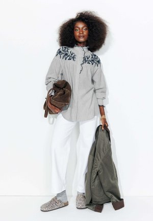 Chemise à rayures avec broderie florale marine, pantalon blanc et mocassins à imprimé animal. Tenant un sac marron et une veste olive.