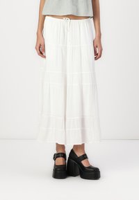 Jupe maxi blanche à volants avec une taille élastique. Tissu léger et fluide avec une texture lisse. Associée à des chaussures à plateforme noires.