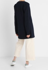 Lång marinblå tunika med rund halsringning, kombinerad med avklippta off-white byxor med fransiga fållar och vita sneakers med gröna detaljer.