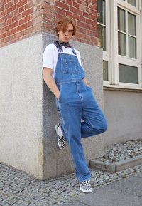 A világoskék farmeroverall állítható pántokkal és elülső zsebekkel rendelkezik. Fehér pólóval és fekete-fehér kockás slip-on cipőkkel párosítva.