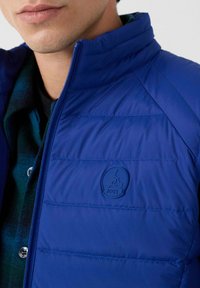 Nahaufnahme einer Person, die eine blaue Steppjacke mit hohem Kragen und ein teilweise sichtbares grün-kariertes Hemd darunter trägt.