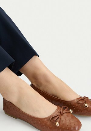 Stopy w brązowych, płaskich baletkach z cienkiego plecionego materiału, ozdobionych małymi kokardkami i złotymi koralikami z przodu, w połączeniu z ciemnoniebieskimi spodniami.