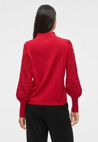 Pull rouge avec un col montant, présentant des manches ballon tricotées avec un motif en diamant. Le corps lisse et les manches texturées créent un contraste.