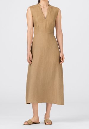 Robe midi beige sans manches avec encolure en V, portée par une femme debout, associée à des sandales plates tressées beiges sur un fond clair.