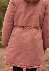 Różowa parka z kapturem, z wkładem w pasie, gładka w texture, z długością sięgającą powyżej kolan. Posiada elastyczne wykończenia i detal z zatrzaskami.