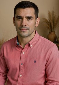 Een man draagt een lichtrode shirt met knopen en een visschubstructuur, voorzien van een donkerblauw logo en een contrasterende gecapitonneerde kraag.