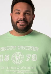Světle zelené bavlněné tričko se bílým potiskem "TROUT TROPHY", grafikou a kulatým logem. Kulatý výstřih a krátké rukávy.