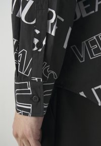 Svart skjorta med djärva vita textmönster. Knäppte manschetter med en mjuk tygtextur. Snygg design med ett modernt, grafiskt utseende.