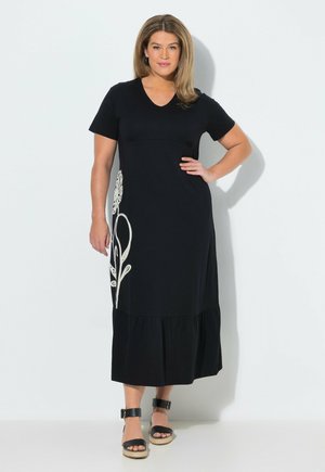 Kvinde med lyst brunt hår iført en sort midi-kjole med hvidt blomsterdesign på siden og sorte platformsandaler.