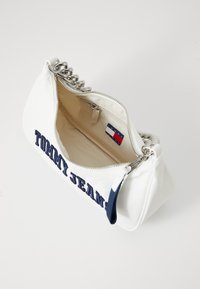 Balta rankinė su jūrinės spalvos "TOMMY JEANS" užrašu, zip uždarymu, sidabriniu grandinės dirželiu ir vidiniais etiketėmis. Lygi medžiaga ir struktūrizuota forma.