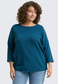 Camiseta de manga larga en color verde azulado con cuello redondo y puños enrollados. Detalle de dobladillo con cordón y tela texturizada. Combinada con jeans de un azul claro.