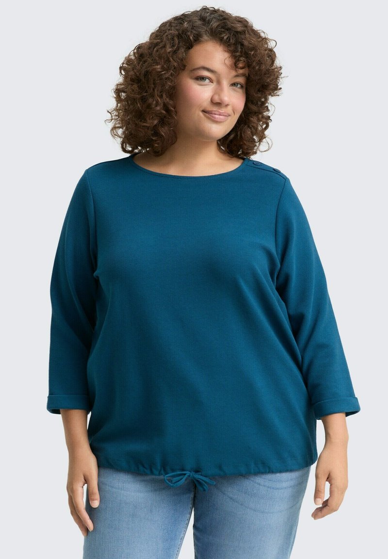 Camiseta de manga larga en color verde azulado con cuello redondo y puños enrollados. Detalle de dobladillo con cordón y tela texturizada. Combinada con jeans de un azul claro.