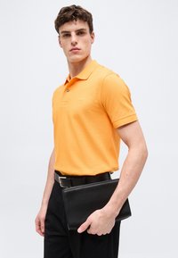 Νεαρός άνδρας με πορτοκαλί πόλο πουκάμισο και μαύρο παντελόνι κρατώντας μαύρο δερμάτινο φάκελο μπροστά σε απλό λευκό φόντο.