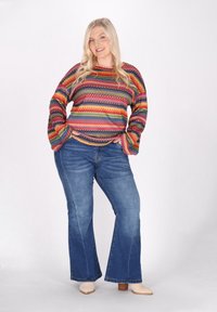 Mehrfarbiger Strickpullover mit langen Ärmeln, der ein Zickzackmuster aufweist. Kombiniert mit blauen Schlagjeans und beigen Ankle Boots.