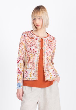 Blondínka v bundě s růžovým, červeným, žlutým a modrým vzorem, přes oranžový top a béžové kalhoty, stojící s jednou rukou v kapse.