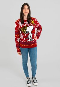Maglione rosso con una grande grafica di Snoopy, fiocchi di neve bianchi e dettagli gialli. Abbinato a jeans blu e sneakers nere.