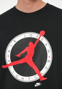 Μαύρο βαμβακερό T-shirt με κόκκι νο και άσπρο γραφικό ενός αναπηδητικού σιλούετ, με την ένδειξη "AIR JORDAN FLIGHT CLUB," και το λογότυπο της Nike στο κάτω μέρος.