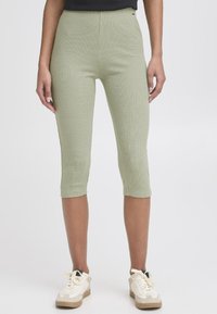Zöld bordás capri leggings, magas derékkal, testhezálló szabással és rövid hosszal, világos színű sportcipőkkel párosítva.
