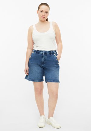 Femme debout, les mains dans les poches, portant un débardeur blanc, un short en jean bleu avec des boutons et des baskets blanches sur fond blanc.