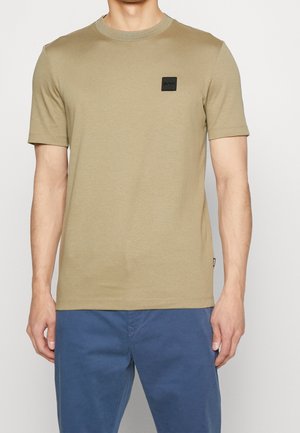 Mann trägt ein schlichtes beige Kurzarm-T-Shirt mit einem kleinen schwarzen Logo-Patch auf der linken Brust und blaue Hose.