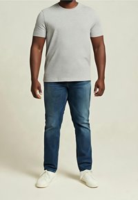 Homme portant un t-shirt gris clair à manches courtes, un jean bleu et des chaussures blanches sans lacets, debout devant un fond uni.