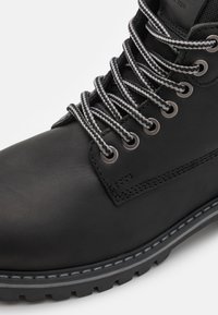 Botín de cuero negro hasta el tobillo con cordones rayados en gris y negro, ojales de metal, cuello acolchado y suela robusta. Textura suave y costura sutil.