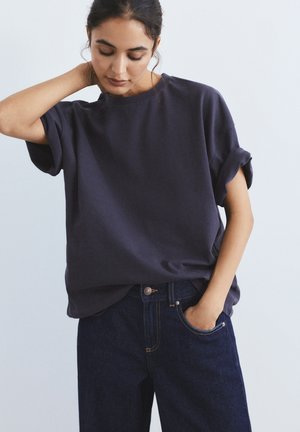 Mladá žena s tmavými vlasmi, oblečená v námornícky modrom tričku s krátkym rukávom a tmavomodrých džínsach, s jednou rukou v vrecku, pozerajúca sa dolu a s rukou na krku.