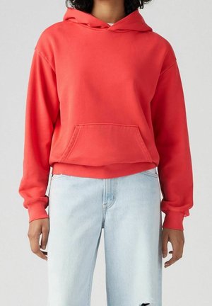 Persoon draagt een fel koraalkleurige capuchontrui met een zak aan de voorkant en lichtgewassen gescheurde jeans, staand tegen een effen achtergrond.