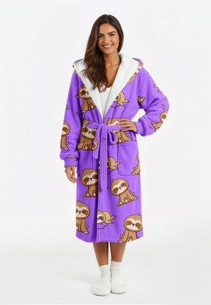 Frau trägt einen lila Fleece-Bademantel mit Kapuze und Faultier-Cartoon-Prints, an der Taille gebunden, kombiniert mit weißen Socken und Hausschuhen.