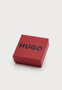 Rød firkantet boks med matt finish, med ordet "HUGO" i fet svart skrift på den øverste overflaten.