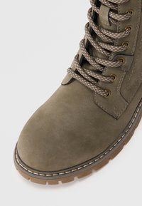 Olivgrüner Schnürstiefel mit einem strukturierten, synthetischen Obermaterial, verstärkter Zehenpartie und kontrastierender brauner Gummisohle. Dicke beigefarbene Schnürsenkel und Nähte.