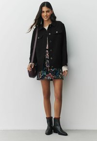 Jachetă neagră texturată cu nasturi argintii, purtată peste o rochie cu model floral. Șosete maro tricotate, cizme negre până la gleznă și o geantă violet.