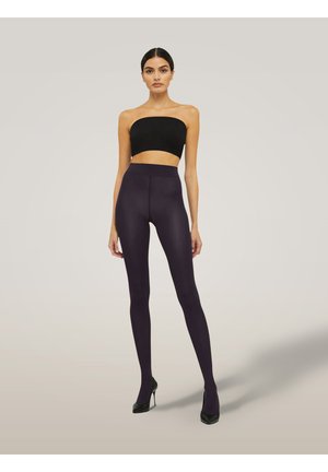 Eine Frau in einem schwarzen trägerlosen Crop-Top, hochgeschnittenen dunkelvioletten Leggings und schwarzen High Heels, die vor einem schlichten Hintergrund steht.