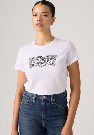 Ung kvinde iført en hvid Levi's t-shirt med et blomstret logo-design og højtaljede blå jeans, stående mod en ensfarvet baggrund.