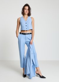 Hellblaues, bauchfreies Weste mit Knopfverschluss, passende hochtaillierte Hose und schwarzer Gürtel. Das Model hält einen blauen Blazer und trägt schwarze High Heels.