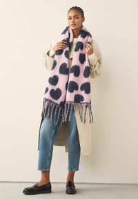 Lyserød uldscarf med marineblå hjertemønstre og frynsedetaljer, parret med en beige frakke og blå jeans. Struktureret og oversized design.