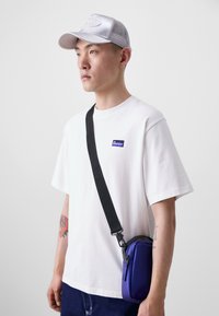 Camiseta branca de algodão com um pequeno rótulo azul, jeans escuros, um boné prateado e uma pequena bolsa transversal roxa com uma alça preta.