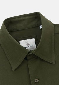 Chemise en coton olive avec un col pointu, fermeture par boutons et une étiquette blanche avec un logo rouge et bleu. Texture douce.