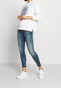 Vit sweatshirt med en blå grafisk design, kombinerad med ljusblåa skinny jeans och vita sneakers. Sweatshirten har sidoflikar och ribbade ärmslut.